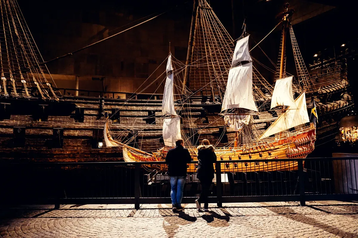 Una exposición en un museo con poca luz en la que se muestra un gran barco histórico de madera con altos mástiles y velas, que recuerda al siglo XVII. Dos visitantes, un hombre y una mujer, están de pie muy juntos, observando los intrincados detalles de una maqueta de barco más pequeña y de colores vivos que se exhibe en primer plano. La escena evoca una sensación de asombro y fascinación por la historia marítima, resaltando el rico patrimonio cultural que se conserva en el museo.