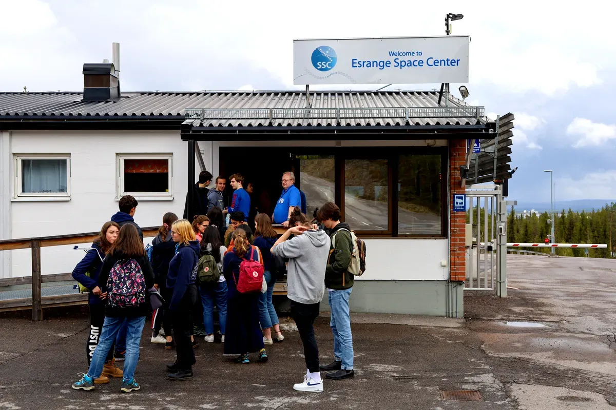 Groupe de touristes rassemblés à l'entrée du centre spatial d'Esrange, en Suède, se préparant pour une visite guidée, avec un panneau indicateur et un ciel nuageux en arrière-plan.
