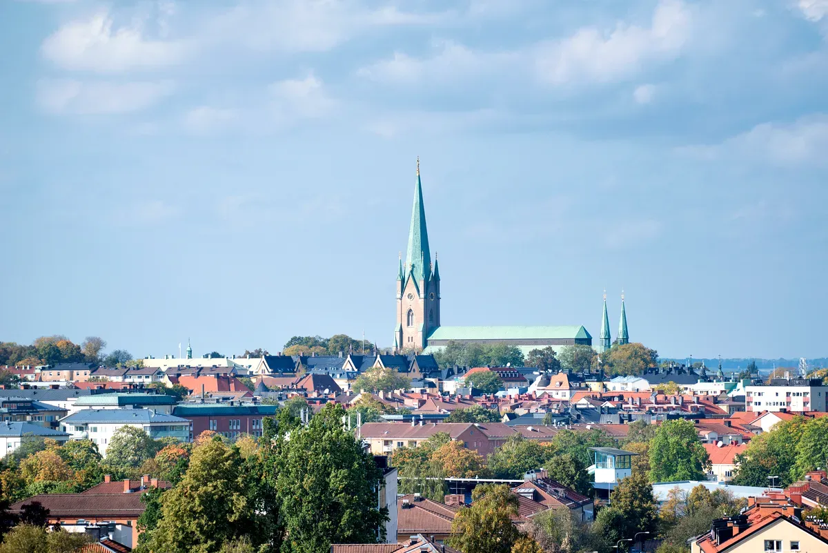 Una vista panoramica di Linköping, in Svezia, con la sua iconica cattedrale che domina lo skyline. L’alta guglia verde della cattedrale svetta sopra gli edifici circostanti, che sono un mix di architettura tradizionale e moderna. I tetti della città, ricoperti di tegole rosse e marroni, si fondono armoniosamente con il verde rigoglioso degli alberi sparsi per il paesaggio urbano. Il cielo luminoso e limpido esalta l’atmosfera serena e pittoresca di questa città storica.