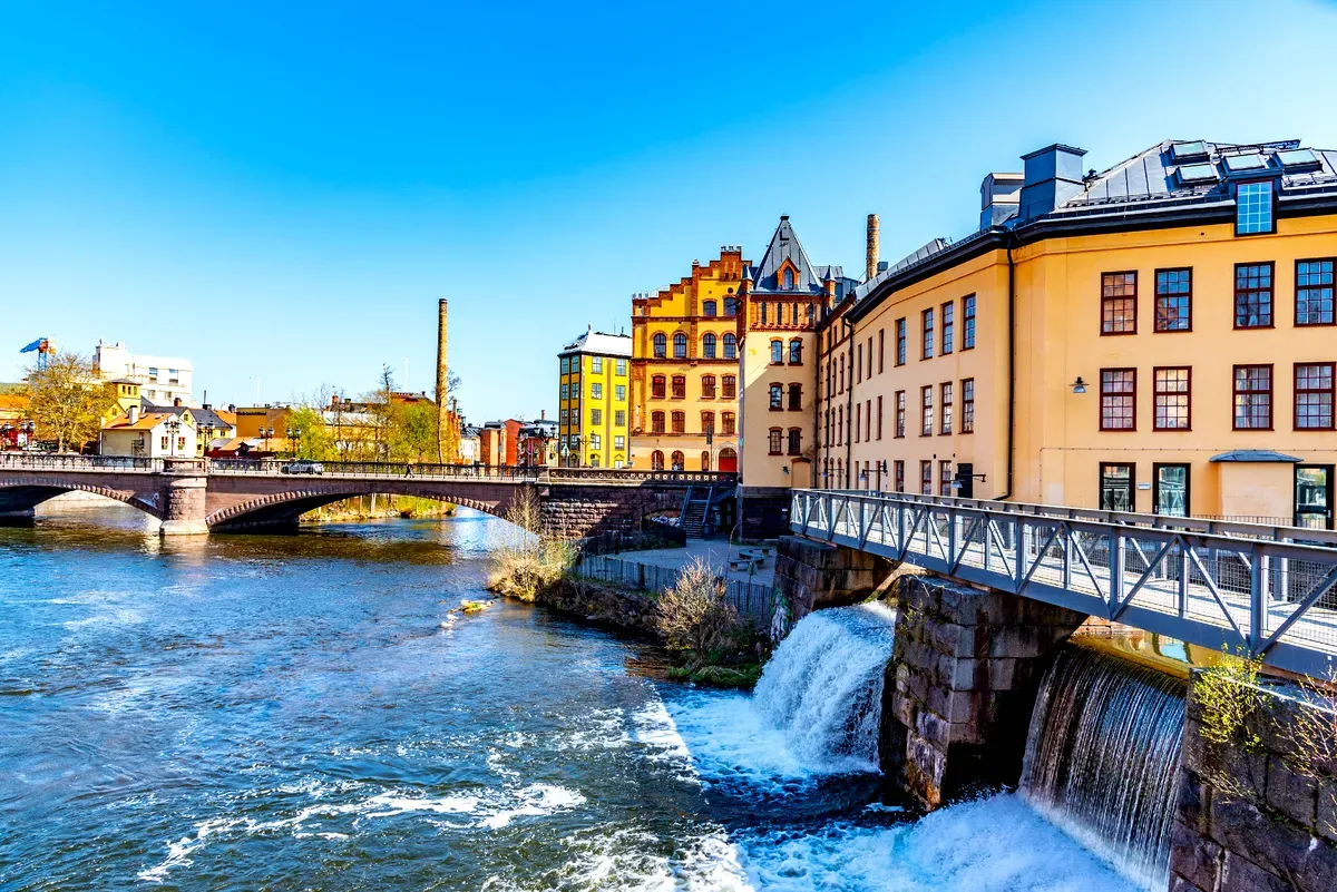 Edifici storici colorati lungo il fiume Motala a Norrköping, in Svezia, con un ponte che attraversa una pittoresca cascata sotto un cielo azzurro e limpido.