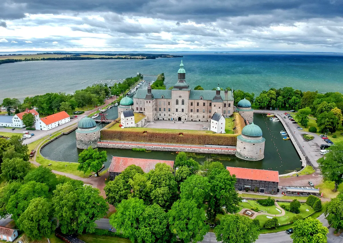 Vista aerea del Castello di Vadstena, un’iconica fortezza rinascimentale in Svezia, circondata da alberi rigogliosi e bagnata dalle calme acque del Lago Vättern. Il castello, con le sue caratteristiche torrette e i tetti a cupola, si erge maestoso all’interno di un fossato, mettendo in risalto il suo storico stile architettonico. Il paesaggio verde e luminoso e il blu vivace del lago sotto un cielo parzialmente nuvoloso creano uno scenario pittoresco e sereno, che riflette il ricco significato storico del castello.