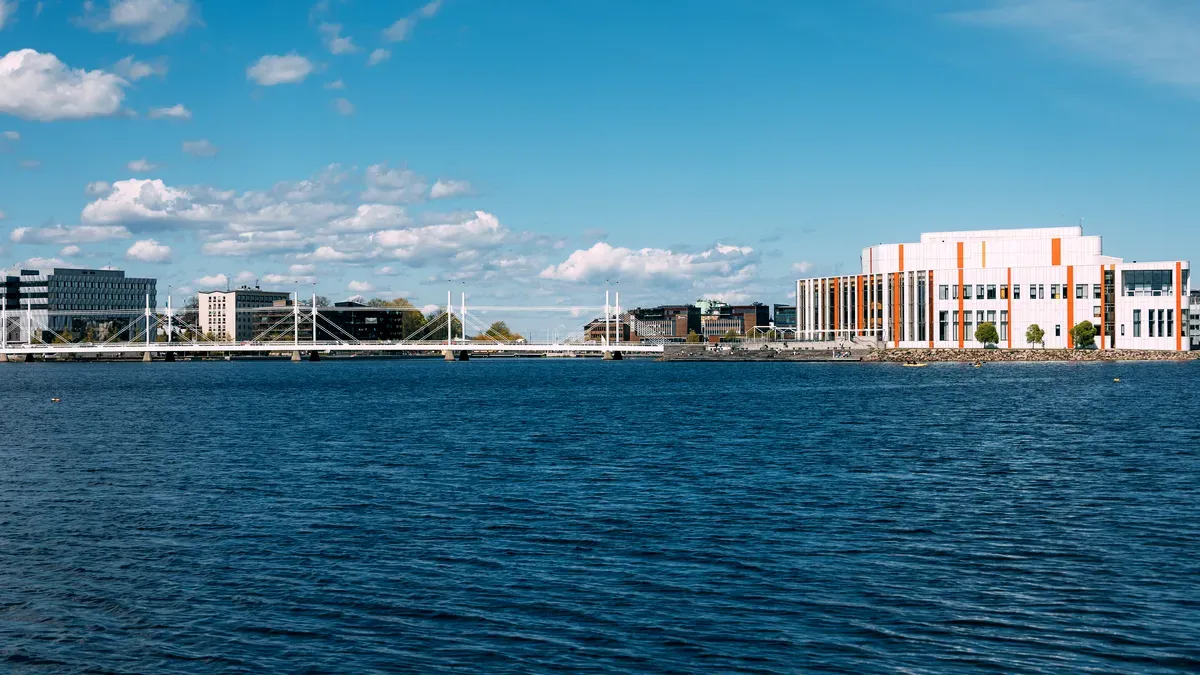 Un moderno panorama urbano visto attraverso un lago tranquillo sotto un cielo azzurro e limpido. Lo skyline è caratterizzato da edifici contemporanei, tra cui una grande struttura bianca e arancione, e un elegante ponte bianco che attraversa l’acqua. I riflessi sull’acqua contribuiscono a creare un’atmosfera serena. Quest’area urbana è circondata da acque tranquille e cieli aperti, offrendo una vista rilassante.
