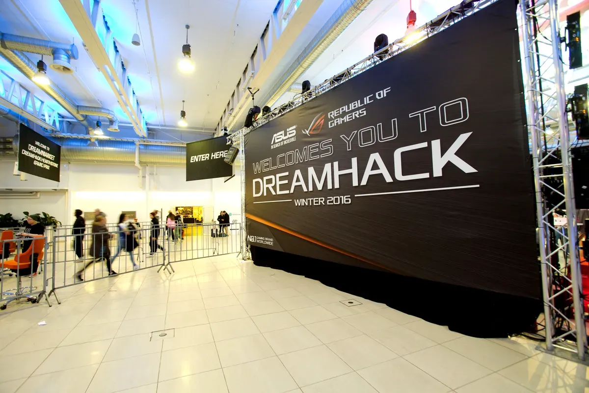 Un grande striscione all’interno di una location moderna e ben illuminata dà il benvenuto ai partecipanti al “DreamHack Winter 2016”, sponsorizzato da ASUS Republic of Gamers. Si vedono persone che attraversano un’area delimitata, dirette verso l’evento. La sede è illuminata da luci intense, ha pavimenti piastrellati puliti e lampadari metallici a soffitto. Lo spazio sembra ben organizzato e pronto per un grande evento dedicato ai videogiochi o alla tecnologia.