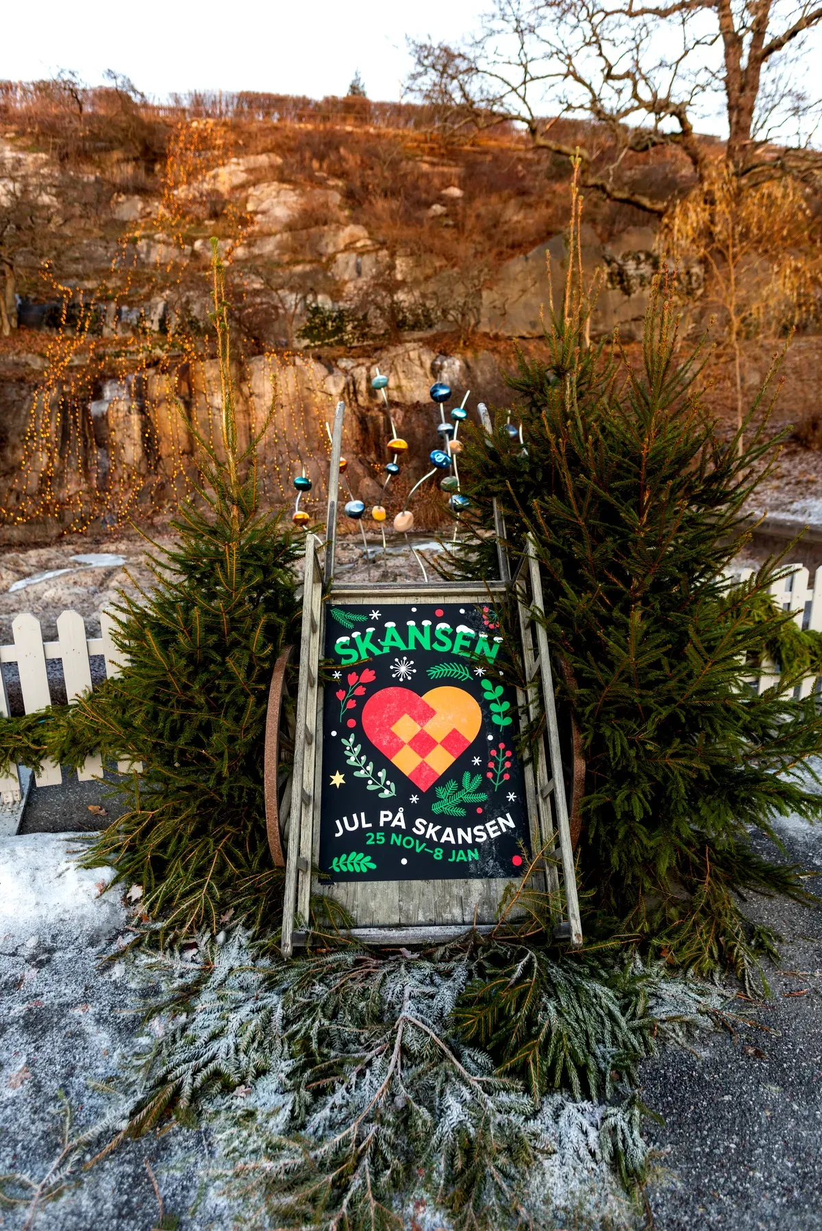 Un cartel festivo con temática navideña en Skansen, colocado en un entorno natural al aire libre con pinos y adornos navideños. El cartel presenta un colorido diseño de un corazón tejido con la palabra «Skansen» en la parte superior y el texto «Navidad en Skansen, del 25 de noviembre al 8 de enero» en la parte inferior. El cartel está rodeado de vegetación invernal y un paisaje cubierto de escarcha, lo que sugiere un ambiente acogedor y propio de esta época del año.