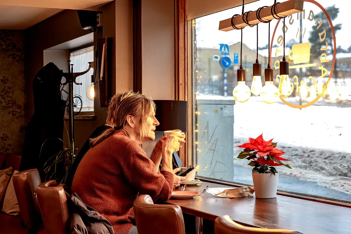 Una mujer sentada junto a la ventana de una acogedora cafetería en Estocolmo, Suecia. Sostiene una taza de café caliente entre las manos mientras mira hacia fuera, con una expresión relajada en el rostro. La cafetería tiene un ambiente cálido y acogedor, con bombillas colgantes y una flor de pascua sobre la mesa. Se ve nieve fuera de la ventana, lo que contribuye al ambiente acogedor de la escena.