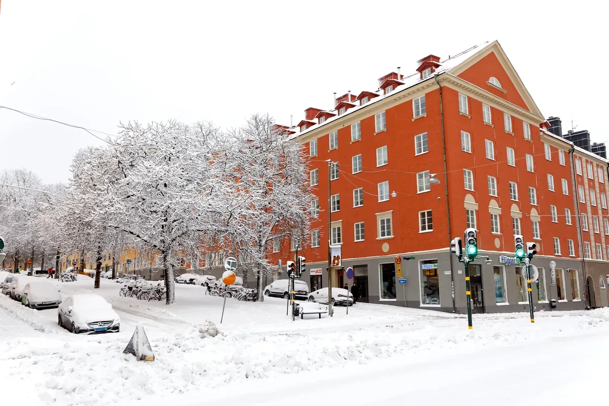 Una calle de Estocolmo cubierta por una gruesa capa de nieve en invierno. La imagen muestra coches, árboles y edificios cubiertos de nieve, con un gran bloque de pisos rojo como punto central. Las ramas de los árboles están muy cargadas de nieve, lo que crea un ambiente tranquilo y invernal. Los peatones caminan por las aceras nevadas y los semáforos están encendidos, lo que indica que la ciudad sigue funcionando a pesar del tiempo.