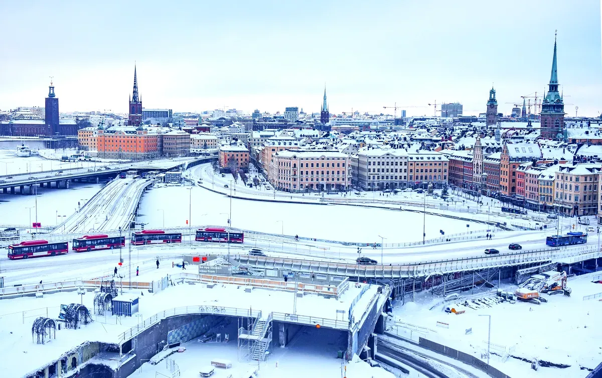 Una vista panorámica de Estocolmo, Suecia, en invierno. La ciudad está cubierta de nieve, con las carreteras y los tejados envueltos en blanco. Se ven varios autobuses rojos en las carreteras nevadas, y al fondo se divisan las emblemáticas agujas de la ciudad, incluida la iglesia de Riddarholmen. Los lagos helados y las calles cubiertas de hielo realzan el ambiente invernal.