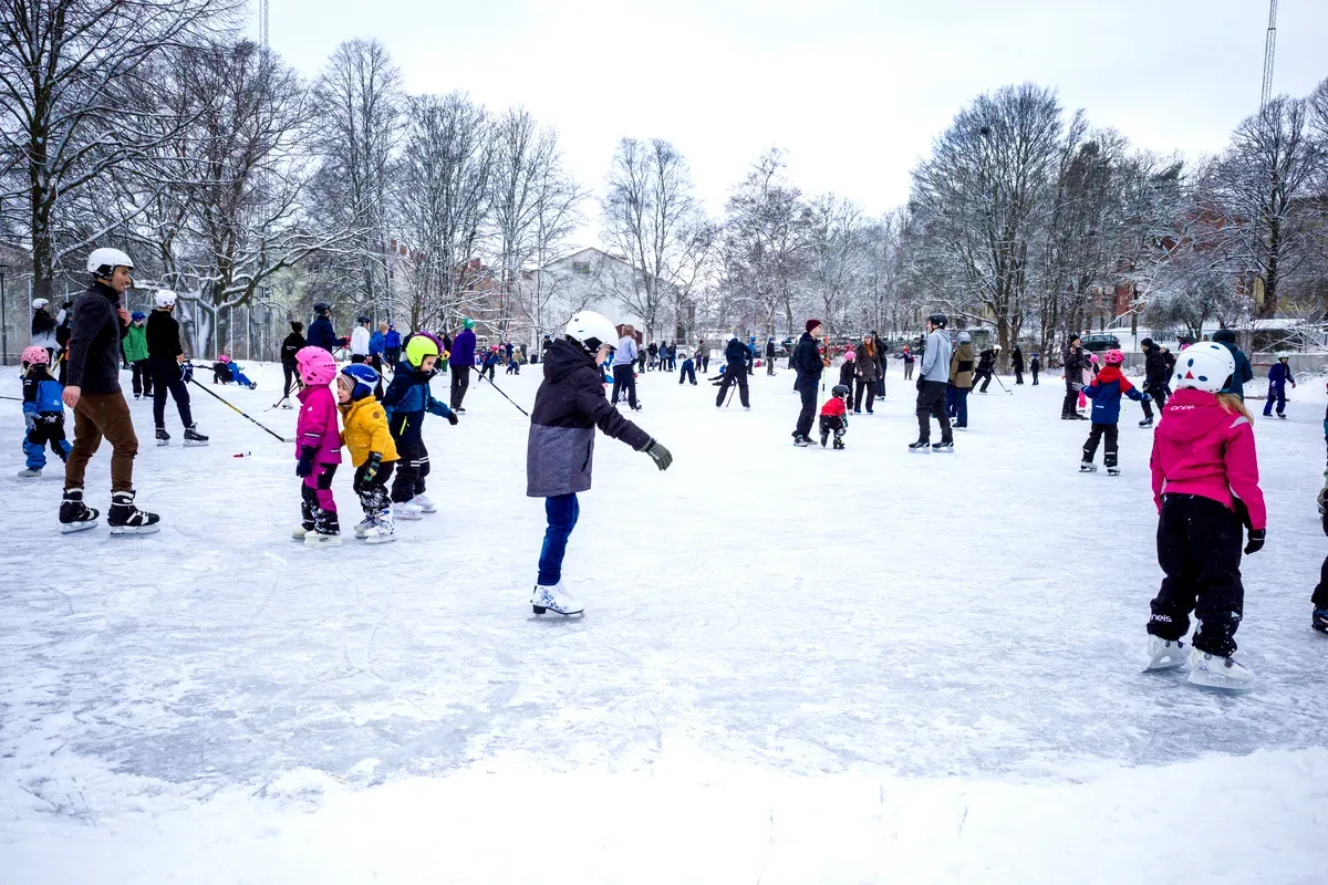 Una animada escena invernal en Estocolmo, donde niños y adultos disfrutan patinando sobre hielo en una pista al aire libre. Todos van bien abrigados con ropa de invierno de colores, y los niños llevan cascos por seguridad. El suelo está cubierto de nieve y los árboles de alrededor están desnudos, lo que contribuye a crear un ambiente invernal. Se ve a familias y amigos riendo y divirtiéndose mientras patinan.