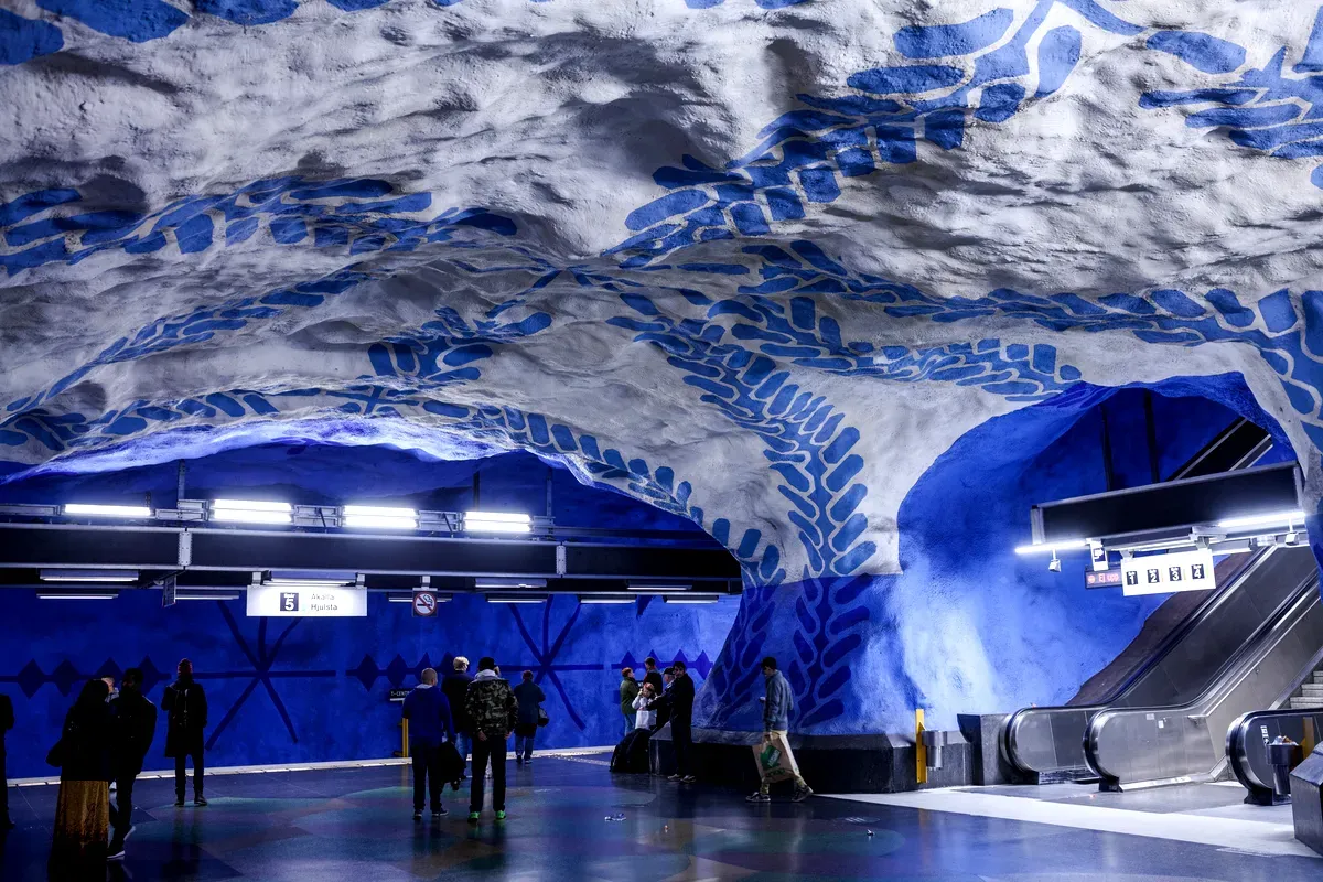 Una foto del interior de una estación de metro de Estocolmo en la Línea Azul, con unas impresionantes paredes tipo cueva pintadas de blanco y azul. Las paredes tienen motivos llamativos que se asemejan a hojas o formas orgánicas, creando un ambiente único y artístico. Los viajeros están de pie y caminando cerca de una escalera mecánica, y la brillante iluminación de la estación añade contraste a los colores vivos.
