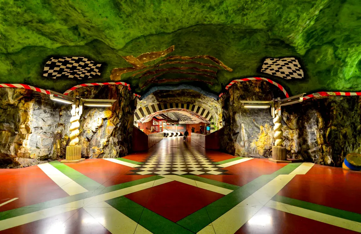Una foto de la estación de Kungsträdgården, en el metro de Estocolmo. Las paredes y el techo están diseñados para parecerse a una cueva, pintados de un llamativo color verde. El suelo tiene un colorido patrón geométrico en rojo, verde, negro y blanco, lo que crea un contraste vibrante. La entrada está flanqueada por pilares de piedra con diseños retorcidos, que conducen a un túnel con motivos a cuadros blancos y negros en el techo. Los detalles en rojo y blanco aportan toques llamativos por toda la estación.