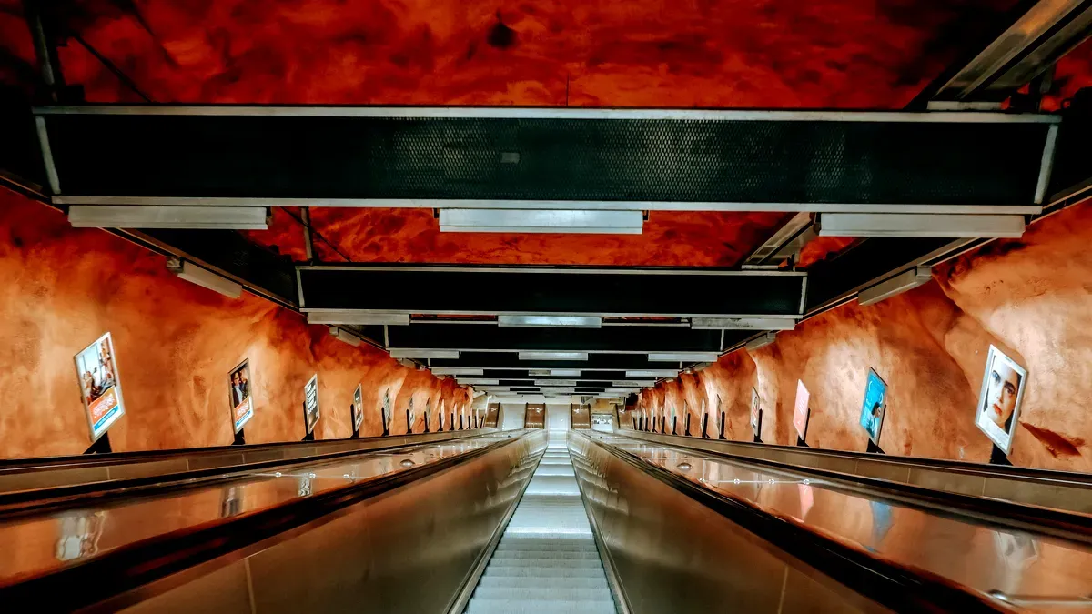 Un largo y espectacular descenso por la escalera mecánica de la estación de metro de Rådhuset, en Estocolmo, Suecia. Las paredes y el techo de la estación están pintados en tonos intensos de marrón rojizo, que recuerdan a una cueva. Las vigas metálicas y las lámparas cuelgan del techo, lo que contribuye a crear una atmósfera industrial y cavernosa. Los anuncios se alinean a ambos lados de la escalera mecánica, que parece extenderse hasta perderse en la lejanía.