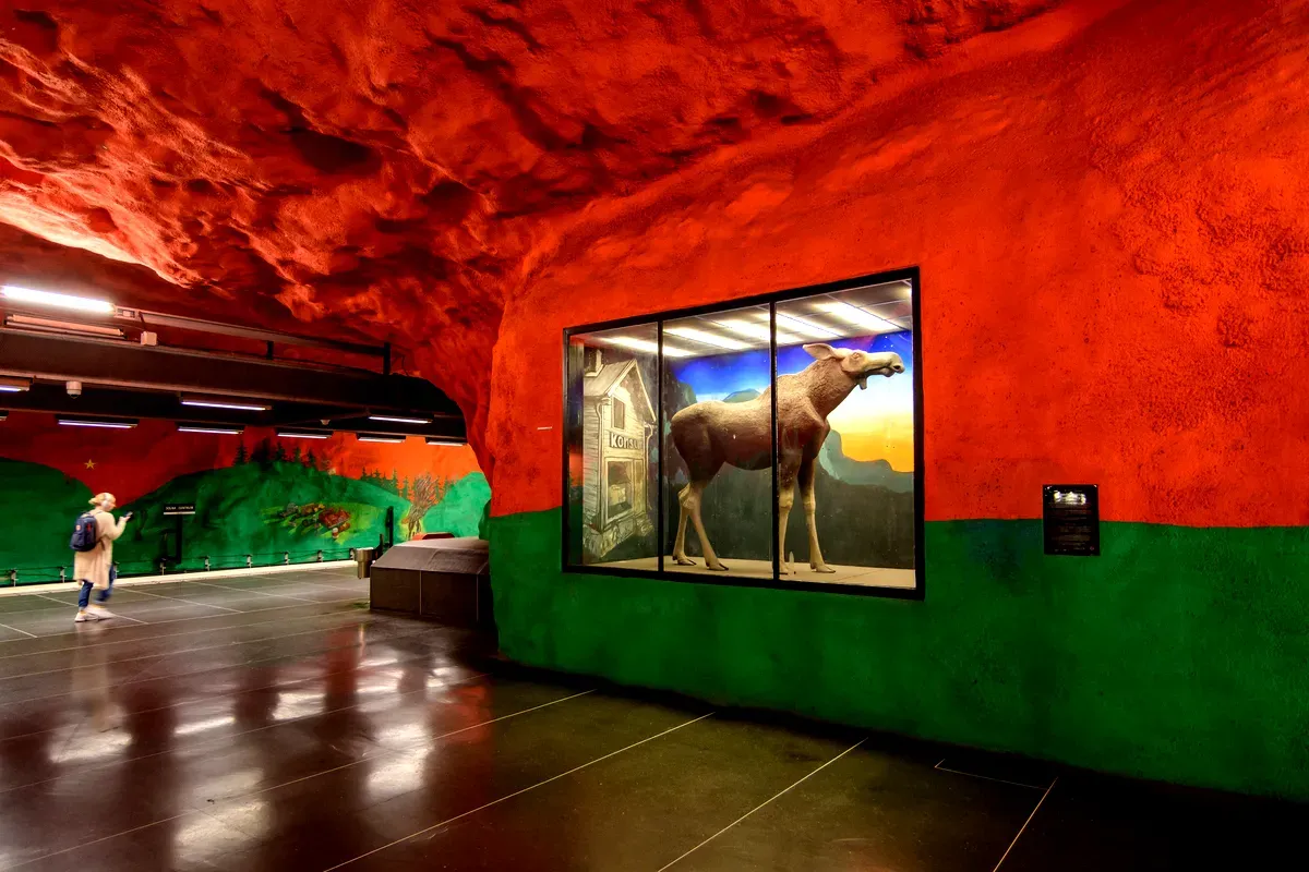 Una colorida instalación de arte urbano en la estación de metro Solna Centrum, en Estocolmo. Las paredes y el techo de la estación están pintados en vivos tonos rojos y verdes, lo que le da un aspecto de cueva. Una vitrina de cristal a la derecha contiene una maqueta a tamaño real de un alce, con un fondo de una casa rural y un bosque, bajo un cielo nocturno estrellado. Se ve a una persona caminando al fondo, a la izquierda, contemplando los vibrantes murales que representan escenas de la naturaleza y paisajes tradicionales suecos.