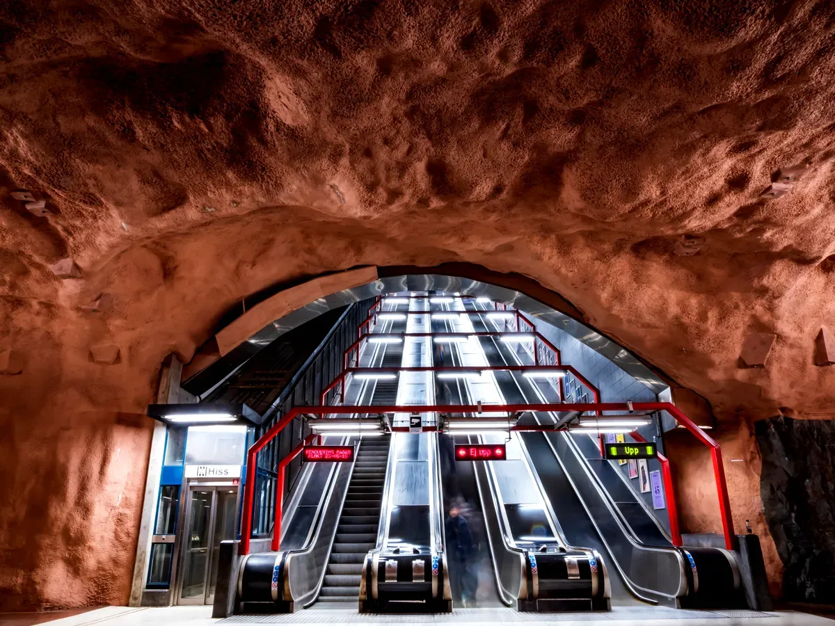 Una foto de la estación de metro Stockholm Stadion, en la que se ven unas escaleras mecánicas que suben a través de un techo con aspecto de cueva, pintado en un tono marrón rojizo intenso. La textura de roca natural de las paredes le da a la estación un aire rústico y orgánico. Las modernas escaleras mecánicas, rodeadas de barandillas metálicas rojas, contrastan marcadamente con el entorno naturalista. Se ve a gente desplazándose por las escaleras mecánicas, y una iluminación brillante ilumina el espacio.