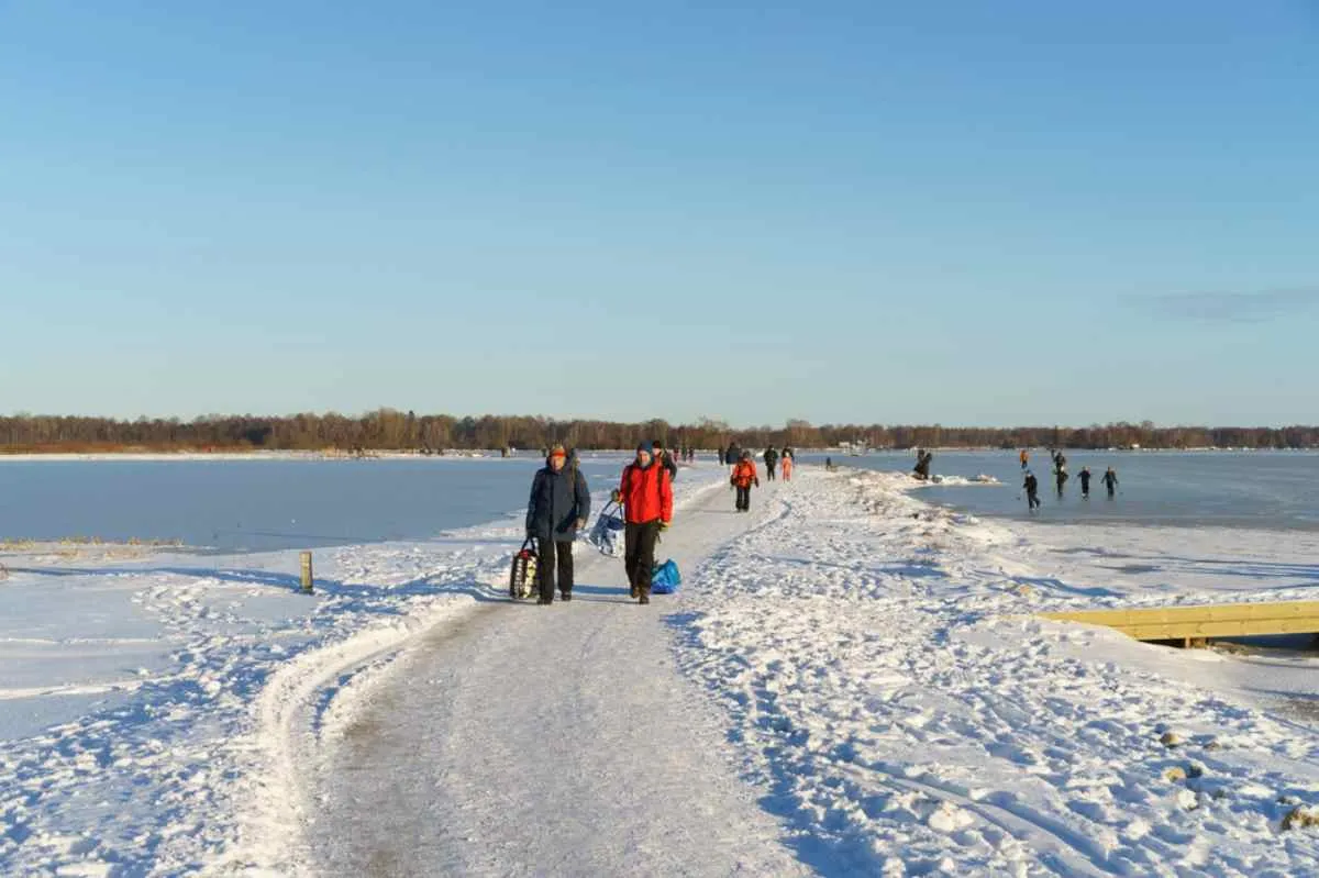 Un grupo de personas, unas en un lago helado y otras en un sendero nevado, disfrutan de un día soleado patinando sobre hielo a las afueras de una ciudad sueca.