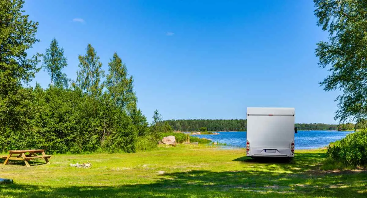 Una autocaravana blanca está aparcada en un paisaje sueco cerca de un lago, en un precioso día soleado en el que la vegetación está en todo su esplendor.