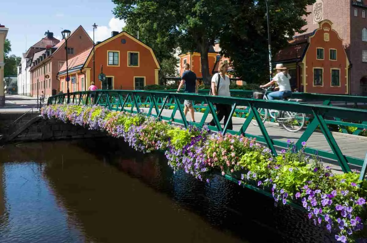 Un groupe de personnes traverse à pied un petit pont dans la ville d’Uppsala, au-dessus d’une rivière entourée de magnifiques fleurs printanières.