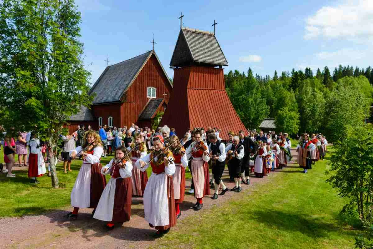 Un groupe de musiciens vêtus de costumes traditionnels suédois apparaît, alignés par trois, à côté d'une église rouge, tandis qu'ils jouent du violon en l'honneur de la fête estivale de la Saint-Jean.