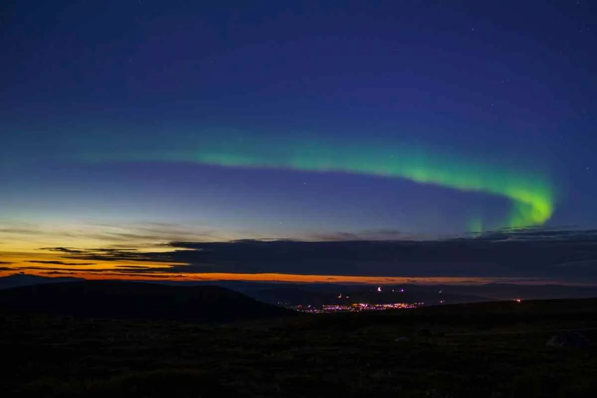Vista general de un paraje natural sueco al atardecer, en la que se ve un pueblo a lo lejos y una tímida aurora boreal en el cielo.