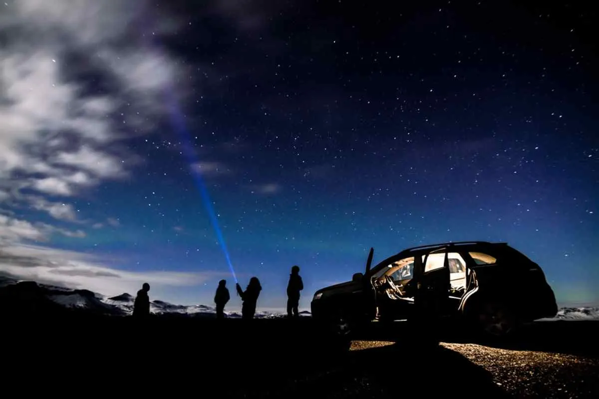 Un grupo de personas a la sombra y bajo un manto de estrellas ilumina el cielo con una linterna y el coche aparcado a su lado.