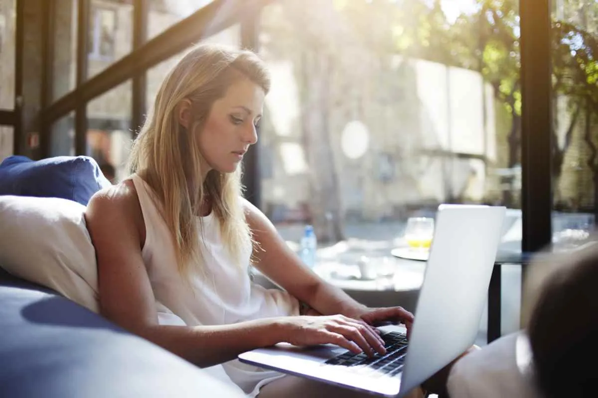 Une femme blonde, dans ce qui semble être un café, navigue sur Internet avec son ordinateur, à côté d’une tasse de café.