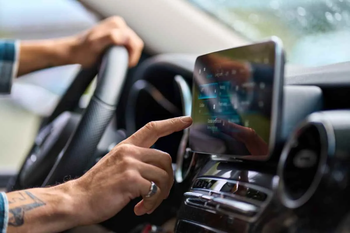 La main d’un homme effleure l’écran d’un téléphone portable posé sur le tableau de bord de la voiture, dans ce qui semble être une tentative de se connecter à une application dédiée aux road trips.