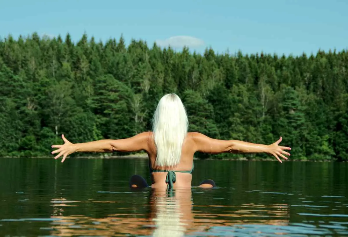 Una donna, con le spalle rivolte verso la riva di un lago svedese in una zona poco profonda, allarga le braccia in un gesto di libertà.