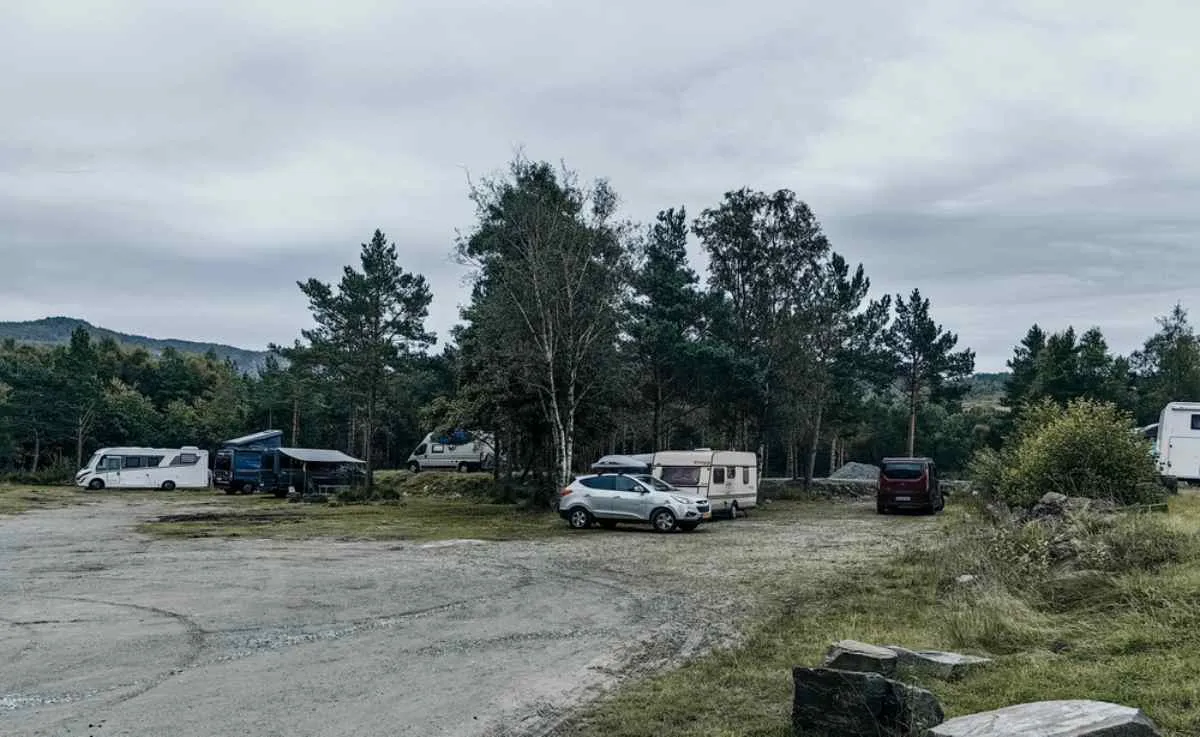 Simple aire de stationnement en gravier accueillant plusieurs camping-cars, fourgons aménagés et voitures, entourée de forêt, illustrant une halte « ställplats » typiquement suédoise.