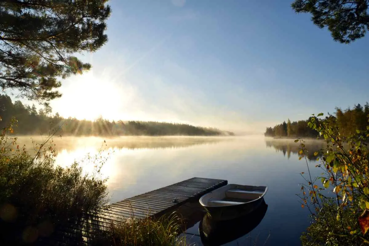 Lever de soleil brumeux sur un lac forestier paisible, avec un petit ponton en bois et une barque : une scène matinale sereine sur un itinéraire en camping-car dans le Småland.