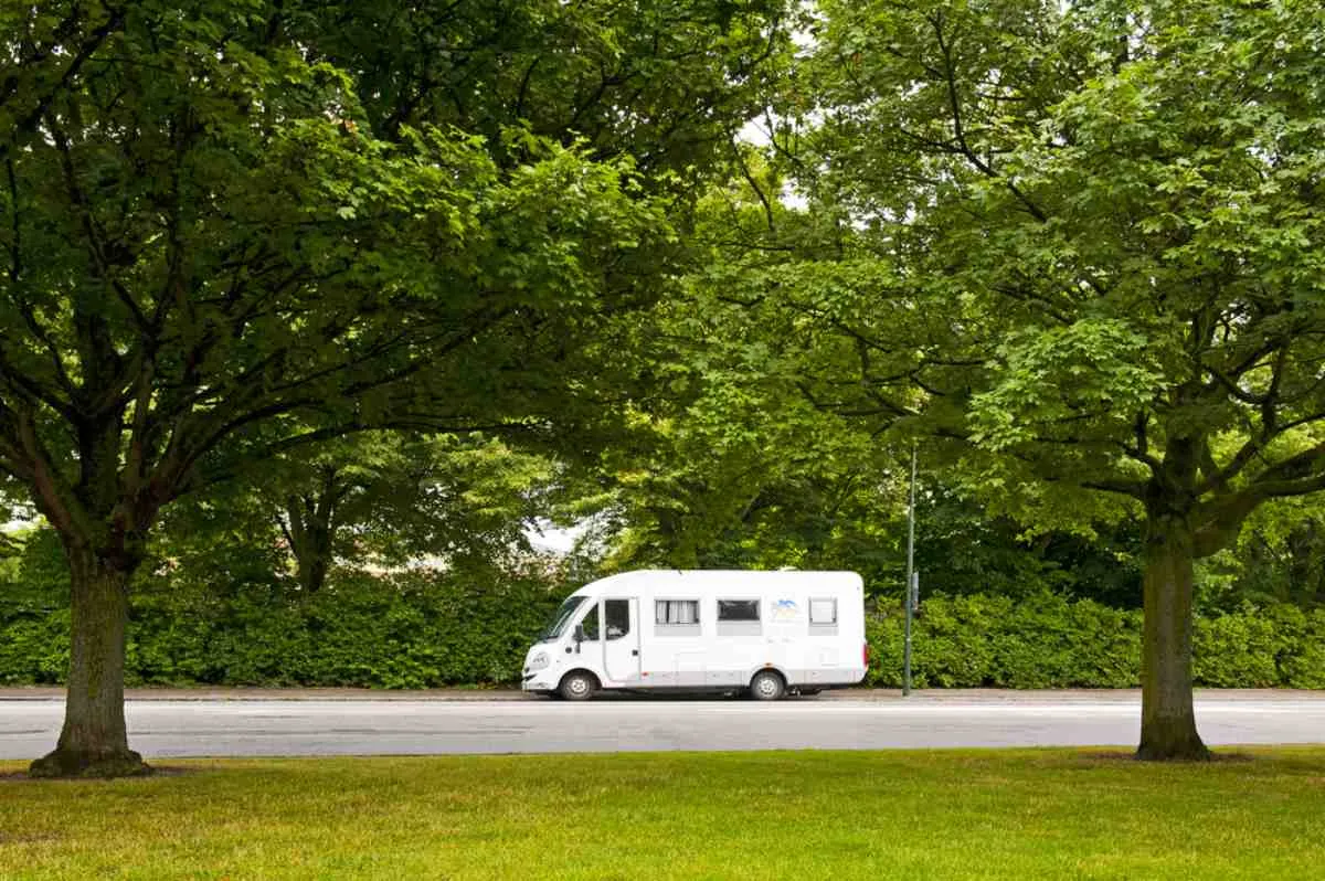 Un camping-car blanc garé sous des arbres au feuillage verdoyant, au bord d’une rue calme du sud de la Suède, illustrant à quel point il est facile de camper au plus près de la nature.