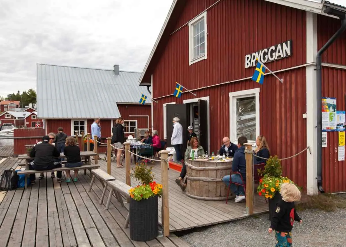 Gente disfrutando de un «fika» frente a la cafetería Bryggan en Bönhamn, sentados en una terraza de madera del puerto rodeada de casitas rojas y banderas suecas.