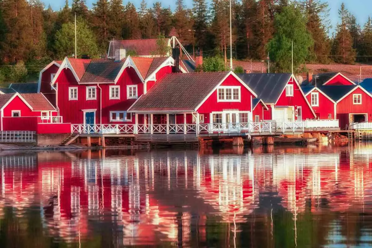 Casas rojas de madera en Norrfällsviken reflejadas en las tranquilas aguas de la Costa Alta de Suecia, rodeadas de bosque bajo la suave luz del atardecer.