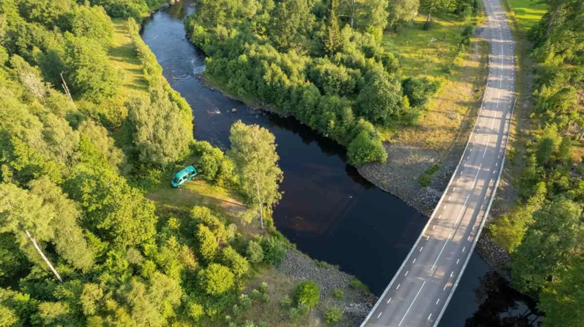 Vista aerea di un camper parcheggiato in riva a un fiume vicino a un ponte, circondato dalla foresta: una sosta panoramica durante un viaggio on the road a Stoccolma.