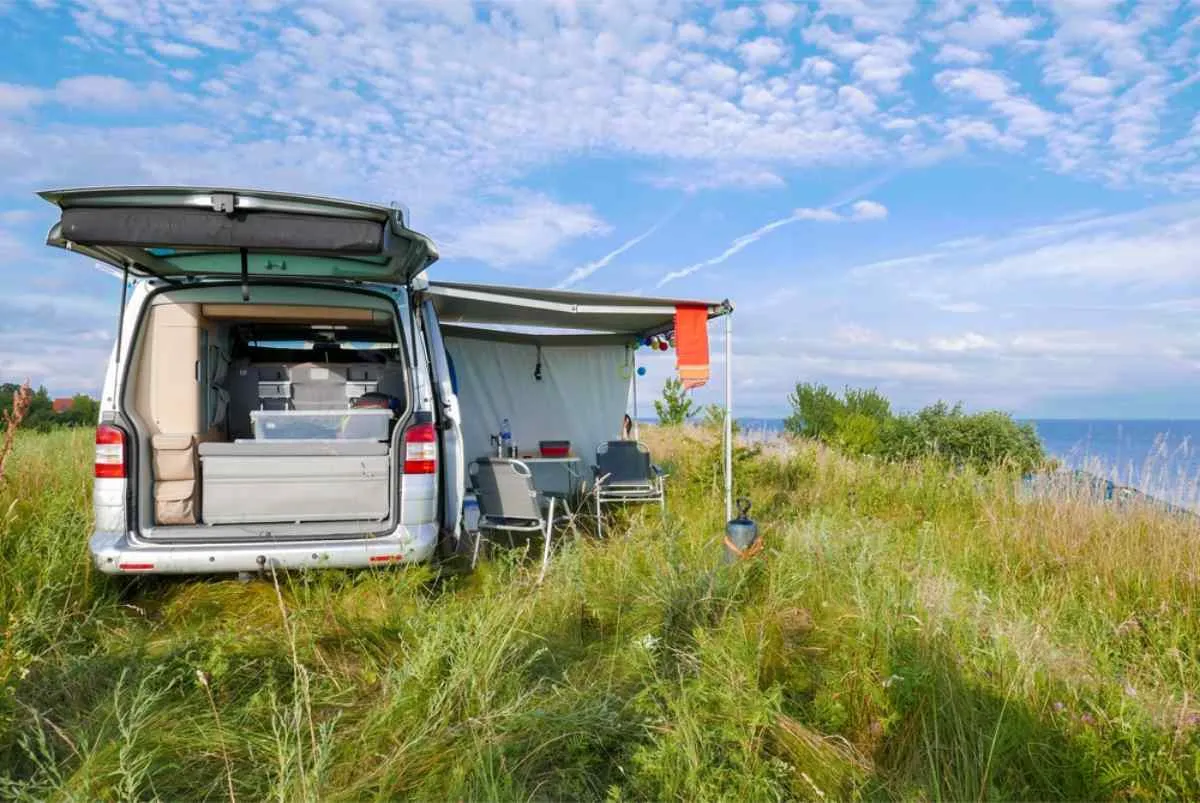 Camping-car garé sur une prairie côtière verdoyante, avec le hayon arrière ouvert et un auvent déployé, ainsi que des chaises et une table au bord de la mer.
