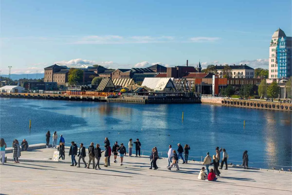 Des personnes se promenant et se détendant sur une promenade moderne au bord de l'eau, avec en toile de fond les bâtiments de la ville et un port paisible, par une journée ensoleillée.