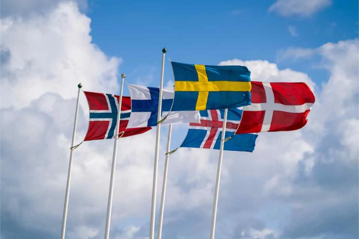Les drapeaux nordiques de la Norvège, de la Finlande, de la Suède, de l'Islande et du Danemark flottent au sommet de hauts mâts, sur fond de ciel lumineux et partiellement nuageux.