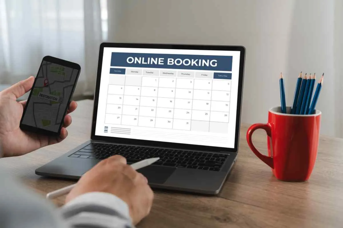 Une personne réservant un voyage en ligne sur le calendrier de son ordinateur portable, tout en tenant un smartphone à la main, assise à un bureau.