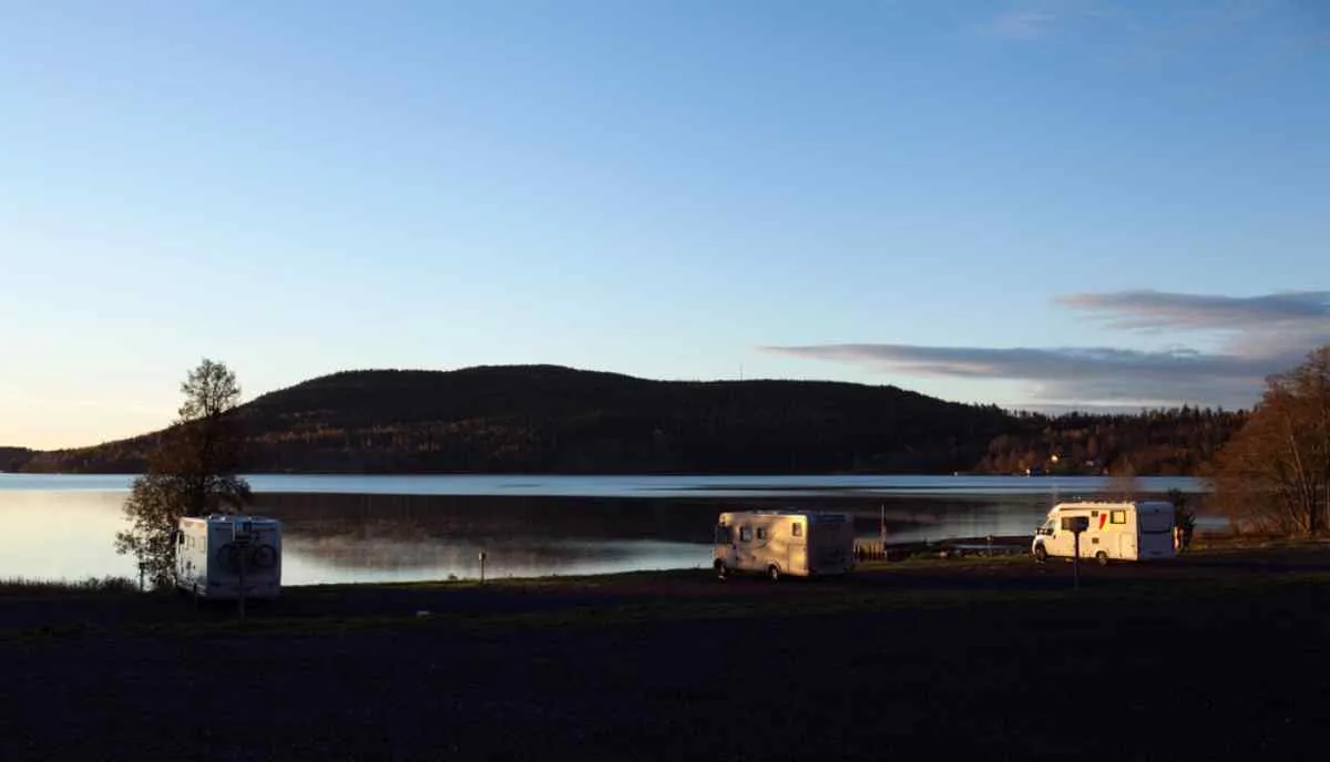 Des camping-cars garés au bord d’un lac paisible au crépuscule, avec en arrière-plan la silhouette sombre d’une colline et un ciel de soirée dégagé.