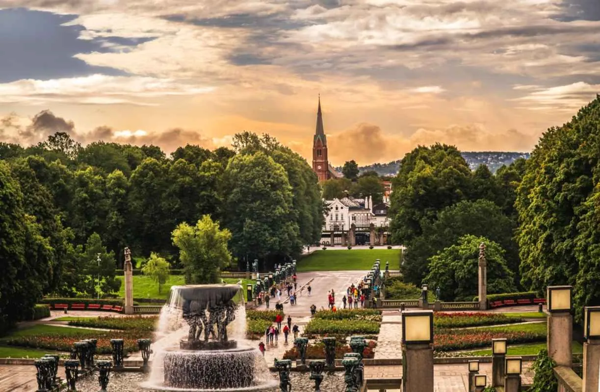 Eine Promenade in Oslo mit einem Springbrunnen im Vordergrund, flanierenden Menschen und einem hohen Kirchturm vor einem dramatischen Sonnenuntergangshimmel.