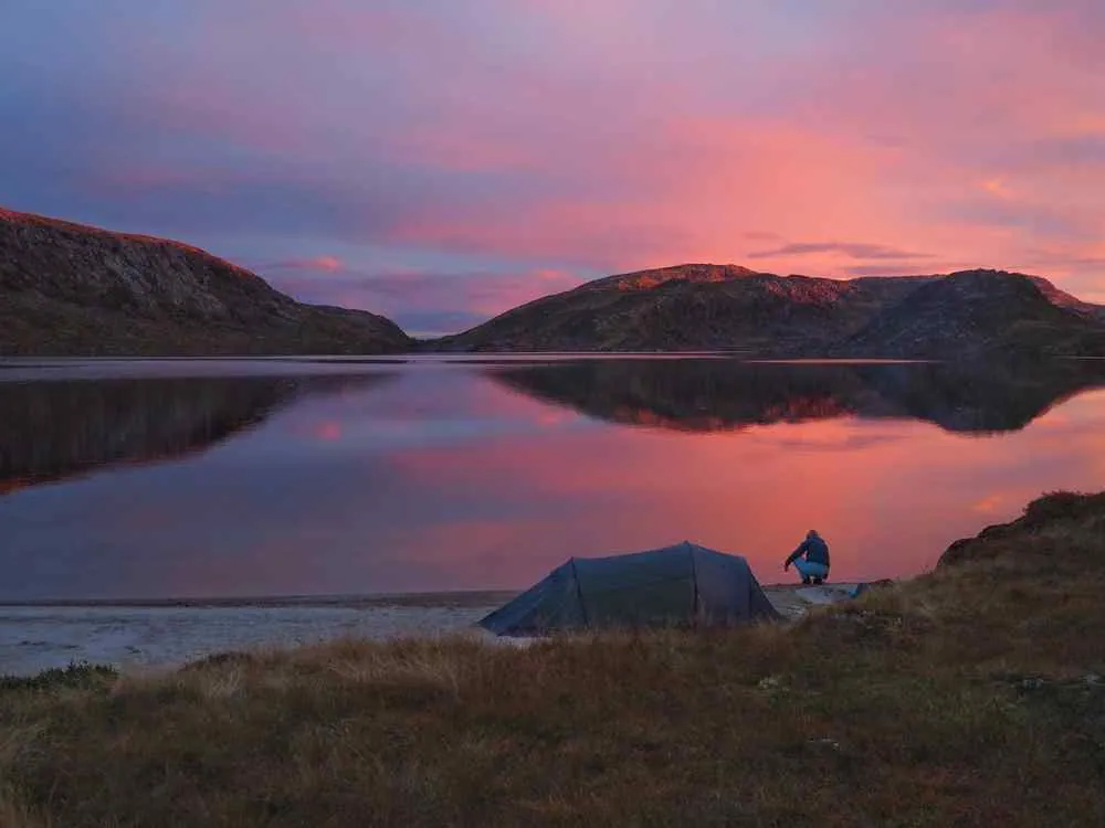 Ein Zelt und ein Campingbus am ruhigen Ufer eines Sees bei rosafarbenem Sonnenuntergang, während sich die Berge im stillen Wasser spiegeln.