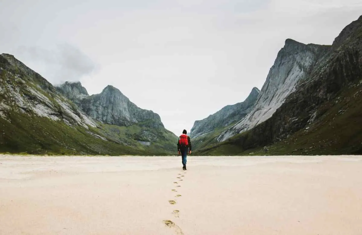 En ensom turgåer med rød ryggsekk krysser en dal med hvit sand mellom de dramatiske fjelltoppene på Lofoten.