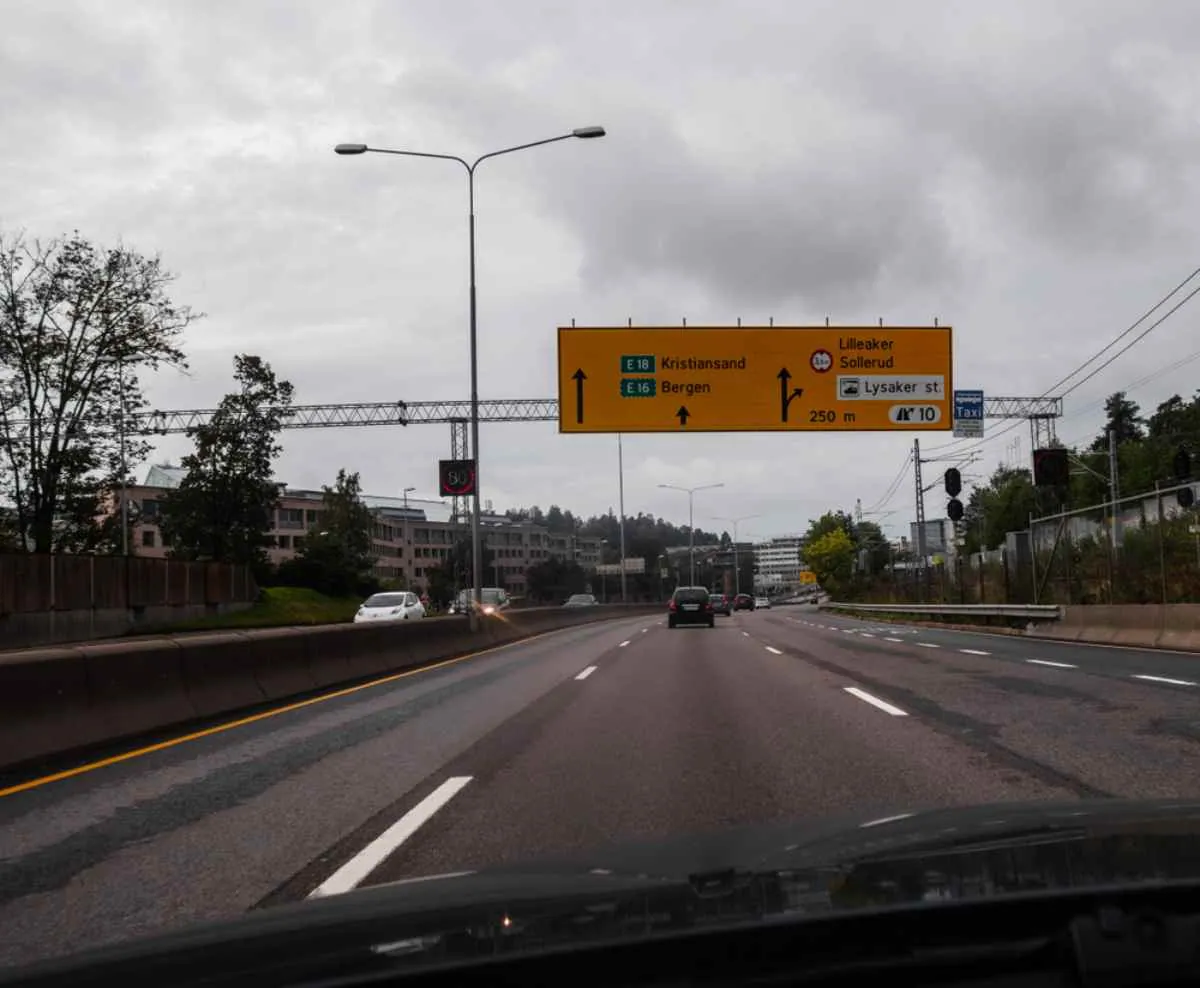 Des voitures roulant sous un ciel nuageux sur l’autoroute E16 près d’Oslo, avec des panneaux routiers jaunes indiquant Bergen et Kristiansand.