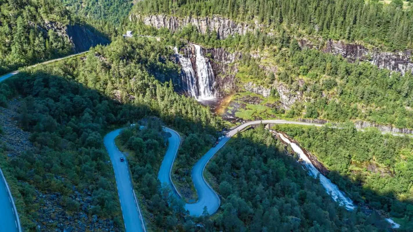 Luftaufnahme eines hohen Wasserfalls neben kurvenreichen Bergstraßen, umgeben von dichtem, grünem Wald.