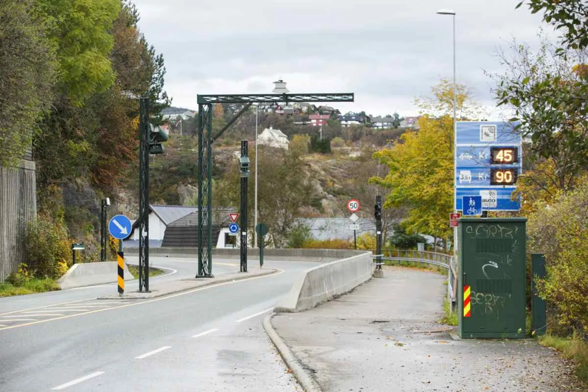 Bomstasjon på landsbygda og prisskilt ved innkjørselen til en bro i Norge, som viser elektroniske bompengesoner og eksempler på priser for passerende kjøretøy.