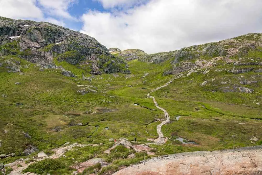 Wanderer folgen einem gewundenen Steinweg durch grüne, felsige Berge unter einem bewölkten Himmel.