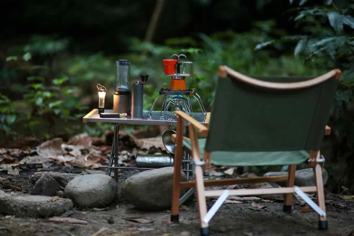 En stol og et mørkegrønt bord står spredt ut i en slags skog, med ulike campinggjenstander på det andre bordet.