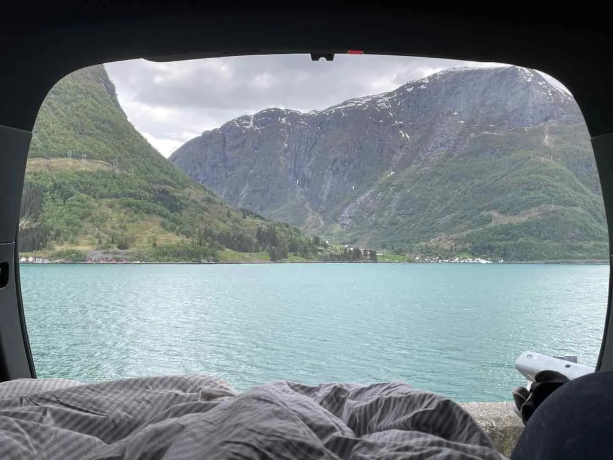Utsikt fra innsiden av en campingbil parkert ved en turkis fjordinnsjø i Norge, med fjell som stiger bratt opp i bakgrunnen.