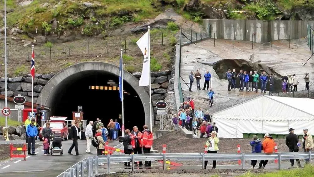 En folkemengde samler seg utenfor inngangen til en fjelltunnel med flagg, et hvitt telt og folk som går på steinete underlag.