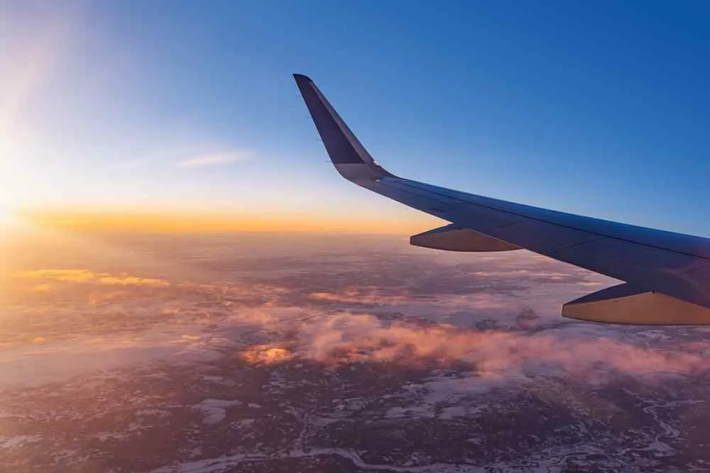 Ein Flugzeugflügel über den Wolken bei Sonnenuntergang, mit orangefarbenem Licht und blauem Himmel über einer dunstigen Landschaft.