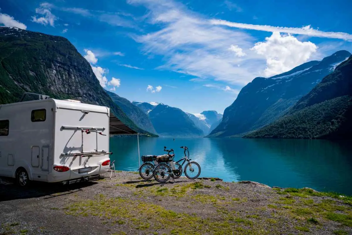 Un camping-car garé au bord d’un magnifique fjord norvégien, avec des vélos et des montagnes se reflétant sur l’eau bleue et calme.