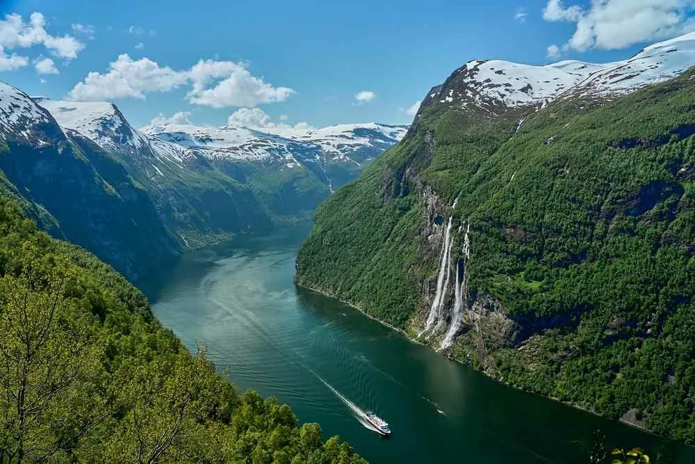 Ein Ausflugsboot auf einem tiefgrünen Fjord zwischen schneebedeckten Bergen, während hohe Wasserfälle unter blauem Himmel die steilen grünen Klippen hinabstürzen.